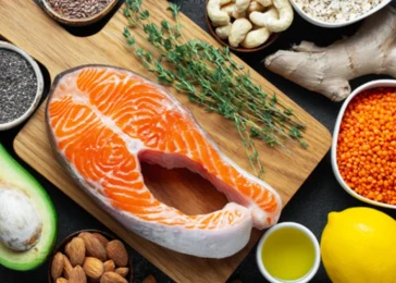 Varios alimentos e cima de uma mesa e uma tabua de cortar carne incluindo peixe, frutas, legumes e temperos representando Quais os alimentos são proibidos para quem tem Doença Renal Crônica